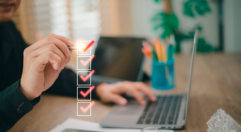 Como Ser Mais Produtivo no Trabalho? Veja dicas, métodos e muito mais! Pessoa dando check em quadradinhos transparentes à frente da imagem. Desfocado, atrás da imagem está um notebook.