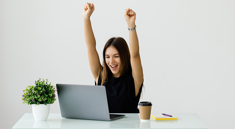Como Ser Mais Produtivo no Trabalho? Veja dicas, métodos e muito mais! Mulher branca comemorando algo em frente a um notebook.