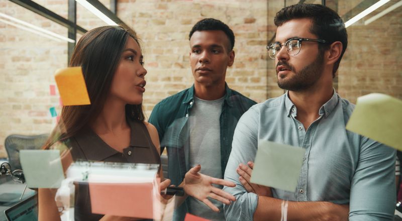 Planejamento Estratégico: Como fazer dicas e exemplos Dois homens e uma mulher debatendo em frente a uma parede transparente com alguns postites grudados.