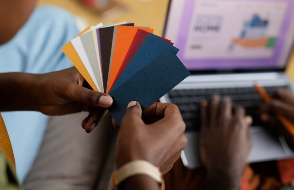 Como criar um logo para minha empresa? Guia completo com todas as etapas que você deve seguir Mãos negras segruando diversos cards com diversas cores cada um em frente a um notebook