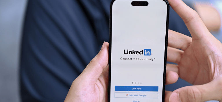 Imagem mostra, de cima, a tela de um celular com a página de entrada do aplicativo do Linkedin, nas mãos de um homem sentado..