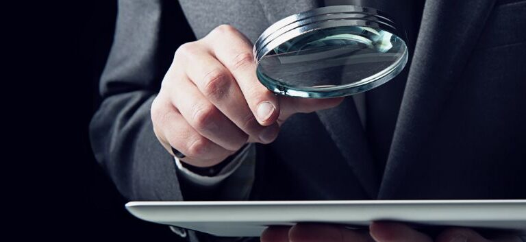 Homem segura uma lupa sobre um tablet.